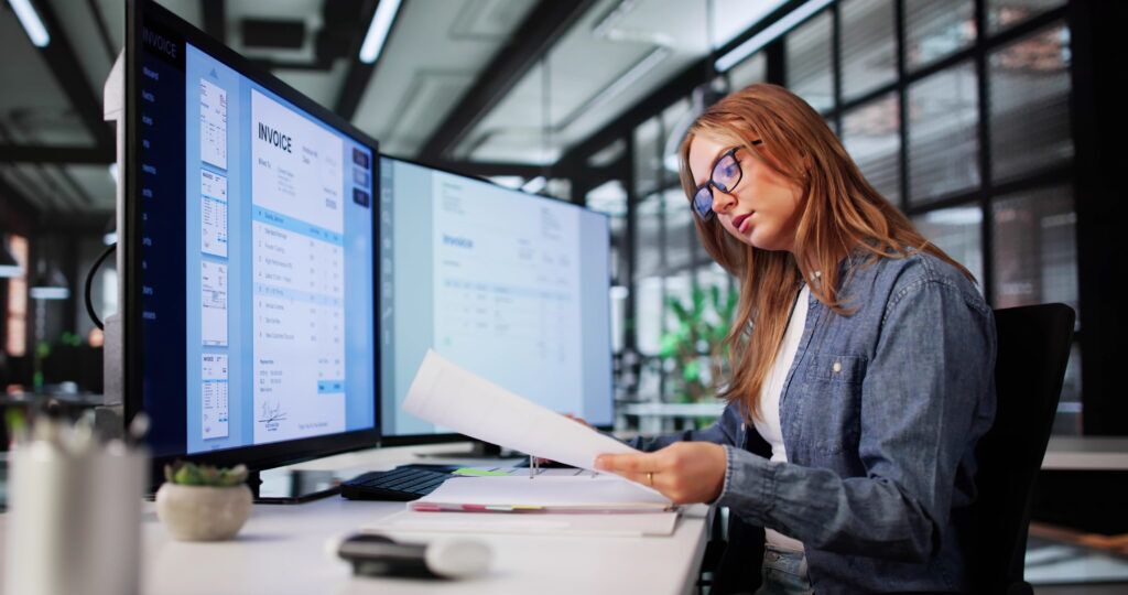 A billing manger compares a printed document to an invoice on a monitor