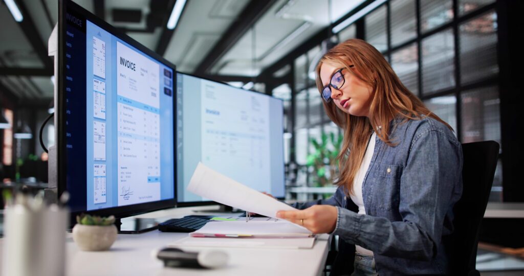 A billing manger compares a printed document to an invoice on a monitor