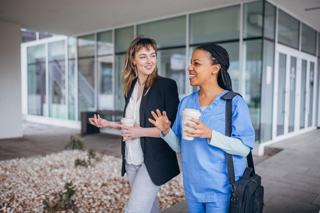 An office manager walks and talks with a nurse. Both or smiling and holding coffee cups.